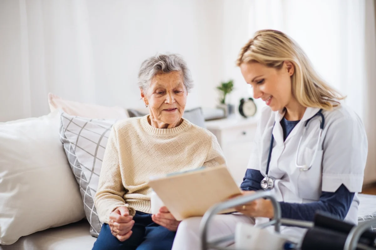 Smiling healthcare professional showing paperwork to elderly woman seated on a sofa, with medical equipment nearby