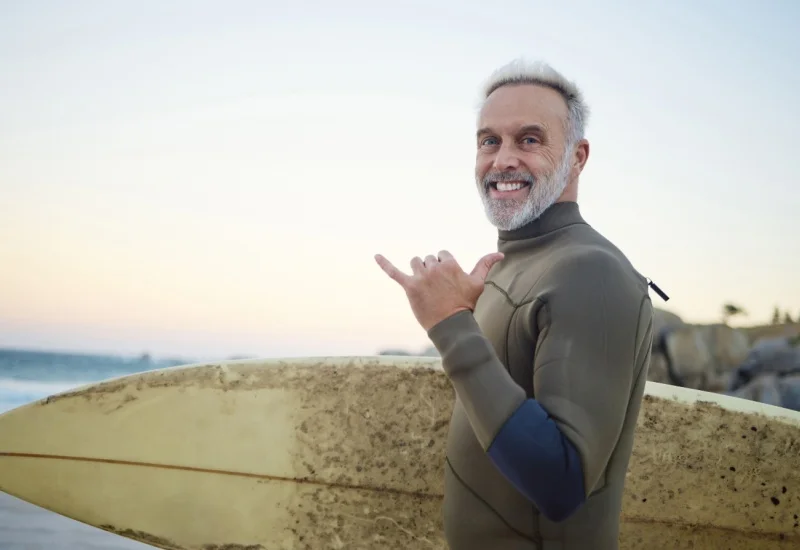 Senior man with surfboard smiling on sunny beach, wearing wetsuit — active lifestyle and summer surfing