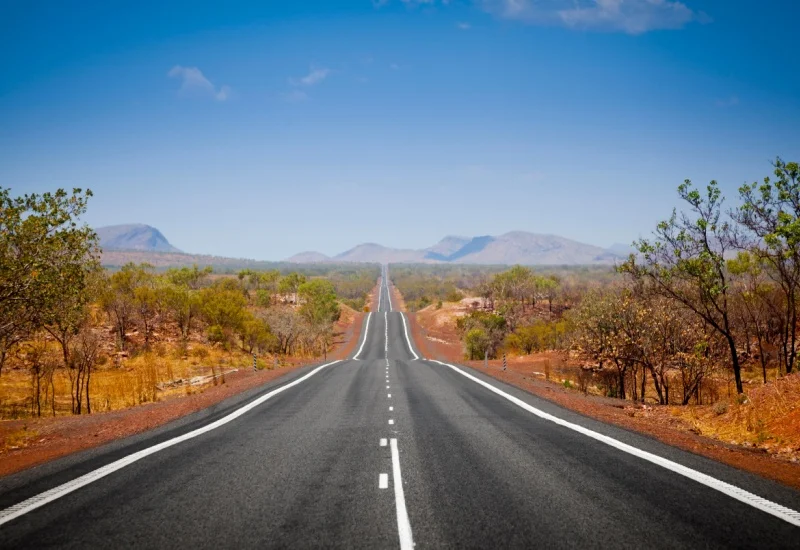 Long straight road stretching into the distance through dry Australian outback landscape with sparse trees and distant hills under a clear blue sky