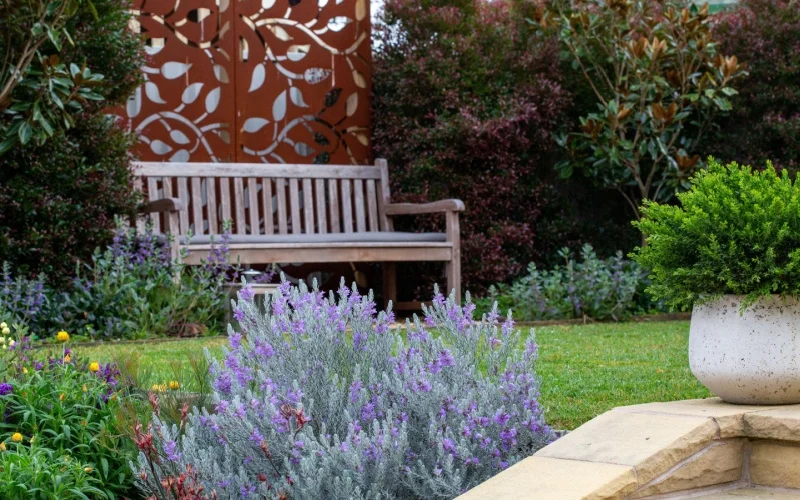 A peaceful garden scene with a wooden bench surrounded by lush greenery, purple flowering shrubs, and a decorative rust-colored metal panel in the background