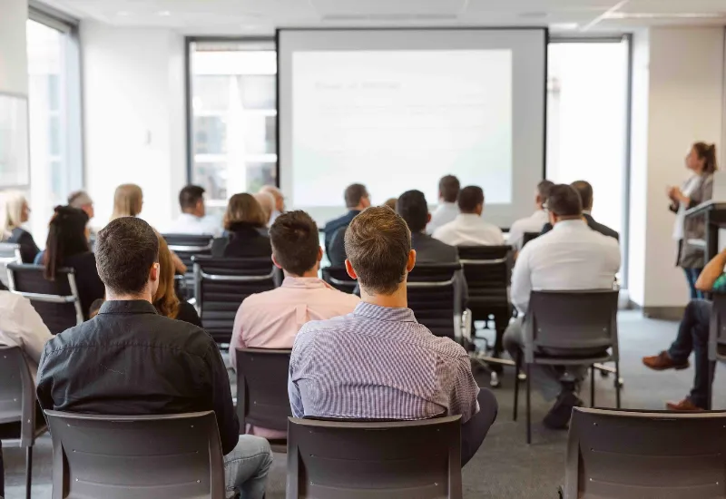 Audience attending a business seminar or corporate training session in a modern conference room, with a presenter speaking in front of a projected screen.